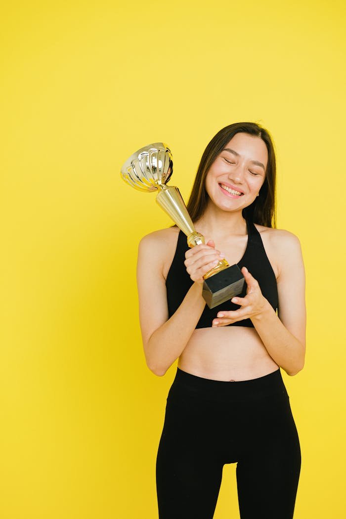 Smiling woman in activewear holding a trophy against a vibrant yellow background, symbolizing achievement.